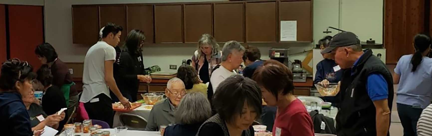 People of several generations making tsukemono (Japanese pickles) together around tables in a church fellowship hall